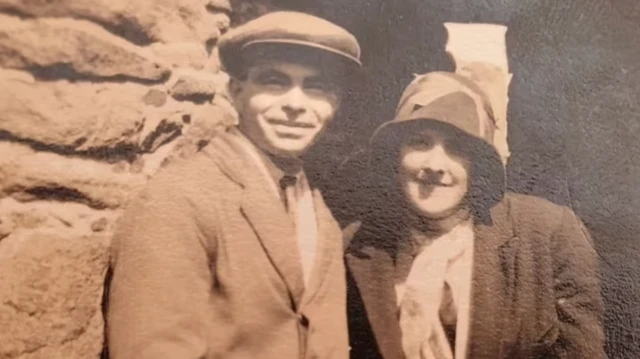John and Phyllis Le Breton pictured smiling in an old sepia photograph. He is wearing a flat cap and suit and she is wearing a hat, coat and scarf. They appear to be standing in front of a stone monument.