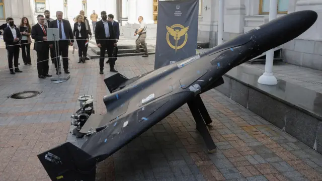 На знімку – дрон «Герань», виставлений як експонат. Він виглядає злегка пошкодженим, а поруч стоїть натовп людей, які його оглядають. На задньому плані видно банер з символікою ГУР