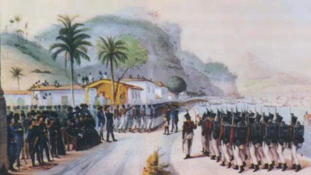 A imagem mostra uma cerimônia militar à beira-mar, com uma tropa de soldados em formação diante de autoridades e civis reunidos em frente a casas coloniais. Ao fundo, destacam-se morros cobertos por vegetação, palmeiras e embarcações ancoradas na baía, compondo uma cena de caráter histórico.
