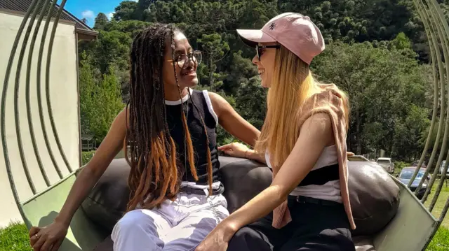 Fotografia colorida mostra duas mulheres sentadas em poltrona ao ar livre, com árvores ao fundo. Elas olham uma para outra e dão risada