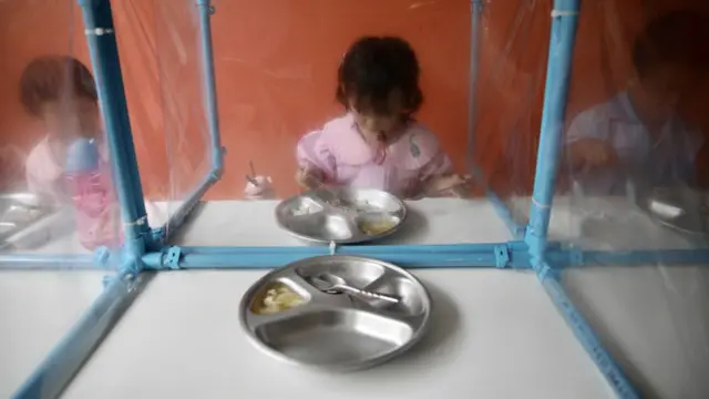 นักเรียนอนุบาลรับประทานอาหารกลางวันโดยมีแผ่นพลาสติกกั้น