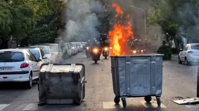 Men in dark uniforms on motorcycles approach a bin burning in the middle of an intersection during a protest