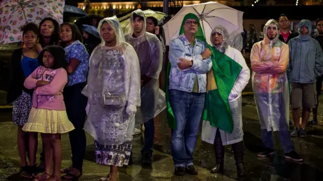 Algunas personas observan los fuegos artificiales desde afuera del estadio con ponchos plásticos y paraguas para protegerse de la lluvia