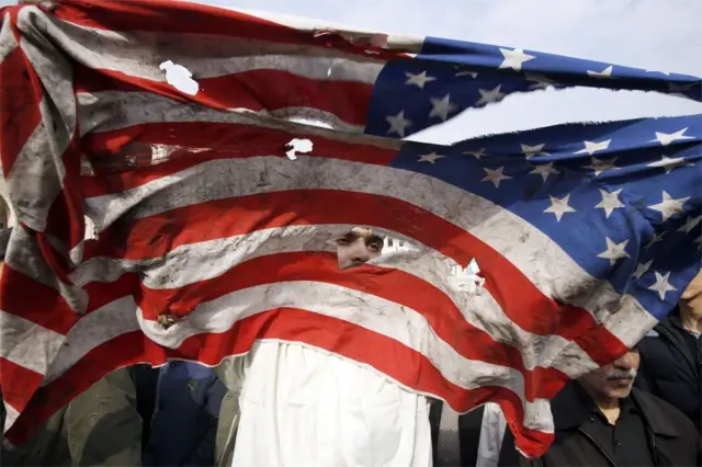 इराकको राजधानी बग्दादमा अमेरिकाले आक्रमण गरेर इरानका वरिष्ठ सैन्य कमान्डर कासिम सुलेमानीलाई मारिदिएपछि तेहरानमा च्यातिएको अमेरिकी झण्डापछाडि देखिएका एक व्यक्ति।