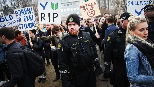 Protestas en Islandia. Policía mira hacia la cámara mientras es rodeado de manifestantes y carteles en oposición al gobierno.