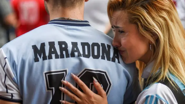 Uma garota chora no ombrojogos de roleta para ganhar dinheiroum homem que veste uma camiseta com o nomejogos de roleta para ganhar dinheiroMaradona