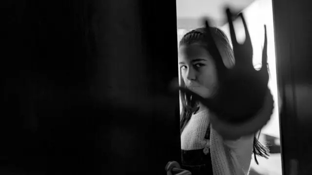 Seorang remaja perempuan berdiri di depan pintu dengan gestur tangan agar berhenti.