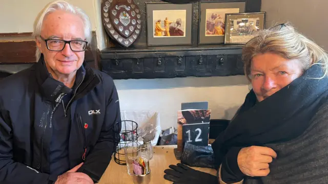 Richard y Carolyn Cater sentados a la mesa. Richard lleva gafas y una chaqueta negra. Carolyn lleva una bufanda.