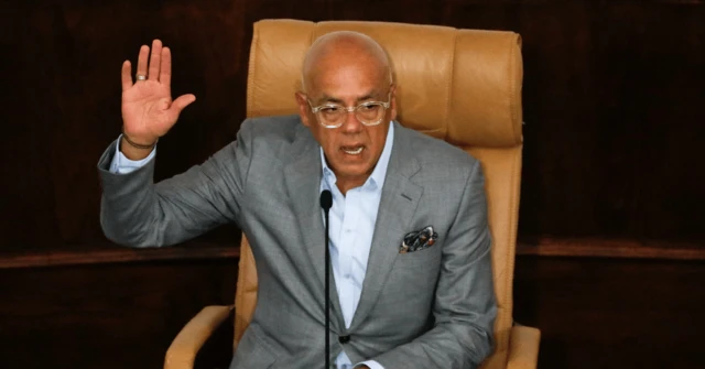 El presidente de la Asamblea de Venezuela, Jorge Rodrñiguez, sentado presidiendo una sesión. Lleva traje claro sin corbata y alza su mano derecha. 