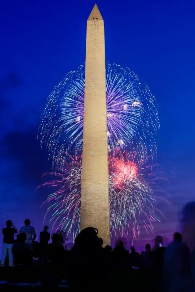 夕焼け空を背に白いワシントン記念塔が立ち、手前に黒い影になった群衆、塔の後ろに赤や青の花火が上がっている