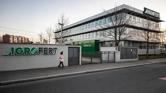 Za murem i bramą stoi biały, trzypiętrowy budynek biurowy z betonu i szkła. Na ścianie widnieje logo Agrofert. Młoda dziewczyna przejeżdża obok muru na skuterze.