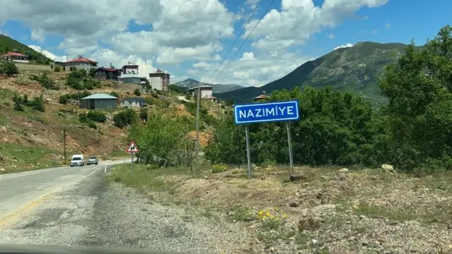 Mavi renkli Nazımiye tabelasının fotoğrafı. İlçe yolu, bazı evler ve arkada yeşil bir tepe görünüyor.