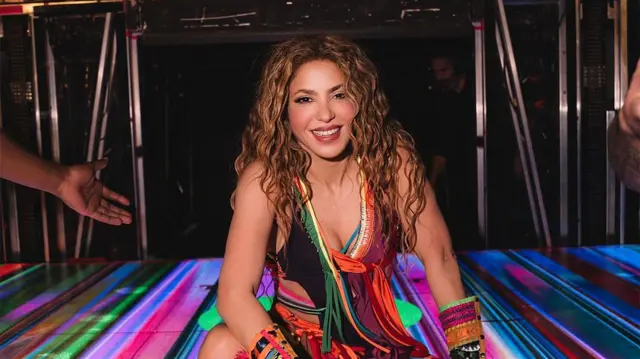 Shakira com uma roupa colorida sentada no palco. Ela sorri para a foto