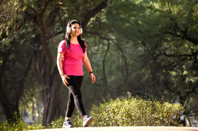 රෝස පැහැති කමිසයක් සහ කළු පැහැති දිග කලිසමක් ඇඳ උද්යානයක ඇවිදින අතරතුර හෙඩ්ෆෝන් භාවිත කරමින් සංගීතයට සවන් දෙන කාන්තාවක්; ඇය ක්රීඩා සපත්තු යුගලක් ද පැළඳ සිටින්නී ය.