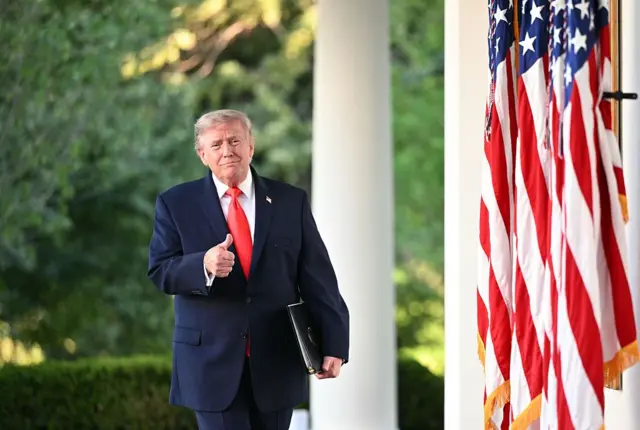 ABD Başkanı Donald Trump, lacivert bir takım elbise ve kırmızı bir kravatla, Beyaz Saray'da üç ABD bayrağının yanında sol koluyla siyah bir dosya tutarken sağ eliyle başparmağını havaya kaldırarak işaret yapıyor.