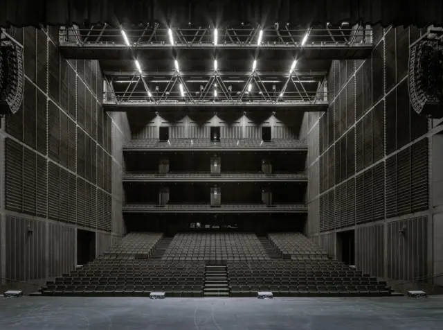 Auditorio amplio visto desde el escenario, con varios balcones, paredes acústicas oscuras y un techo iluminado con estructura metálica.