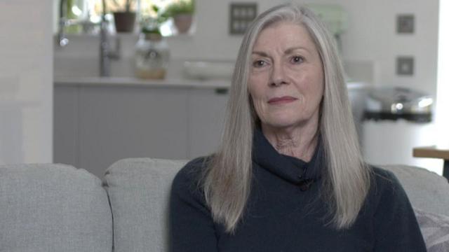 Carolyn Woods olhando para a câmera sentada em um sofá cinza