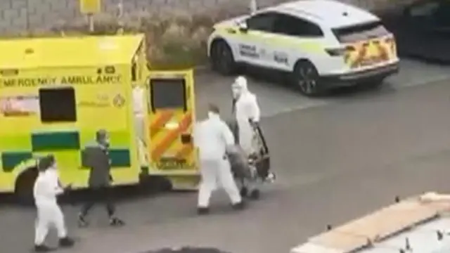 白い防護服を着た3人が、黄色い救急車の前を歩いている。