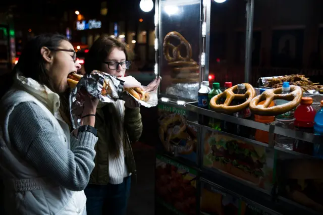 Two girls eat hot dogs outside a fast food stall. It is dark and they are both wearing jackets and jumpers.
