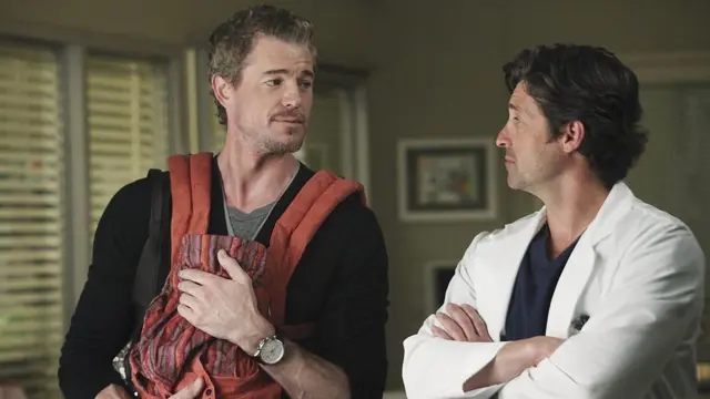 Eric Dane en un hospital; sostiene un porta bebés rojo mientras conversa con el actor Patrick Dempsey que viste bata blanca en una escena de "Anatomía de Grey".