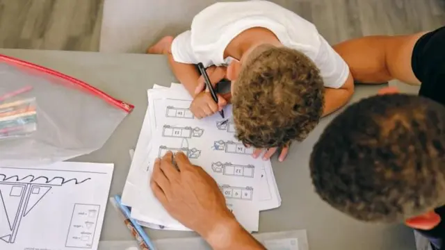 تصویری از بالا، پسربچه‌ای در حال انجام تکالیف مدرسه و پدرش در حال نظارت