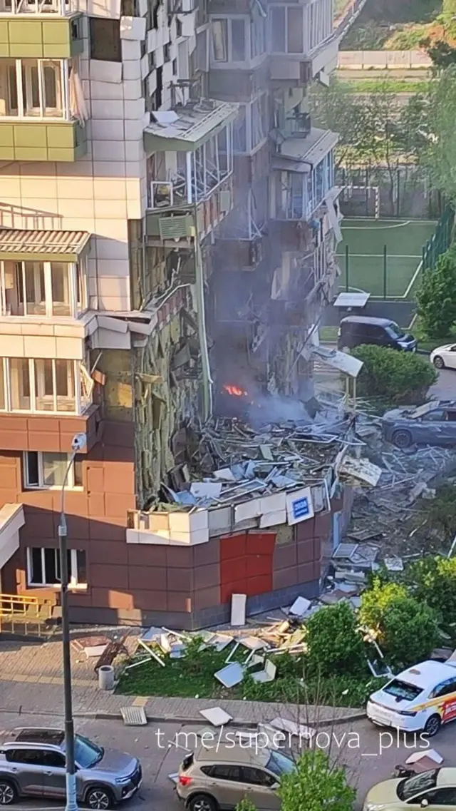 Зовнішня стіна багатоповерхового житлового будинку серйозно пошкоджена — частина нижніх поверхів зруйнована, видно уламки бетону, скла й конструкцій, розкидані навколо. З вікон і балконів в одному місці йде дим, помітне невелике полум’я. Унизу біля будинку стоять припарковані автомобілі, поруч зелена зона й тротуар, також засипані уламками.