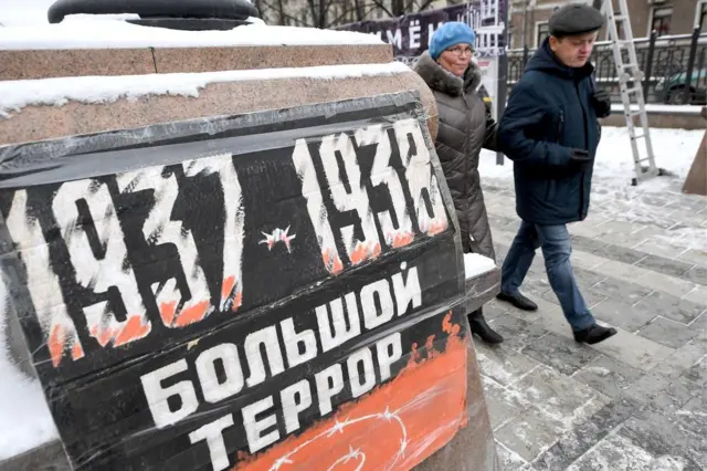 Люди идут мимо камня, на котором установлен плакат с надписью Большой террор