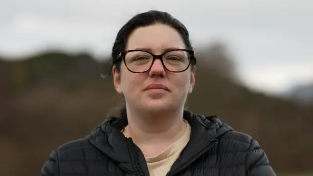 Foto de Tanya, de cabelo preso e óculos, olhando diretamente para a câmera, com a paisagem desfocada de uma montanha ao fundo