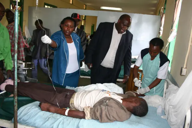 Wahudumu wa afya katika wadi yenye shughuli nyingi nchini Kenya wanamhudumia mgonjwa aliyevaa nguo kamili aliyelazwa kwenye kitanda cha hospitali, huku dripu ya mshipa ikiingia kwenye mkono wake wa kushoto.