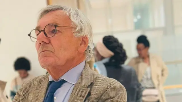 Umugabo wambaye ikote na karavate, inyuma ye hahagaze abagore