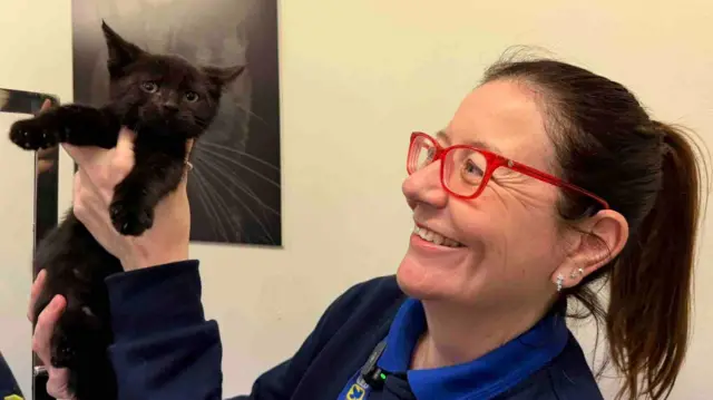 Un gatito negro con las patas al aire, sostenido por una señora con el pelo recogido en una coleta y gafas de montura roja. Lleva un uniforme de la RSPCA y les sonríe a los gatitos.