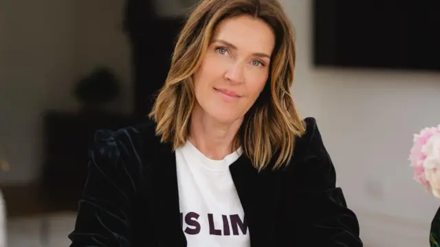 एम्मा बार्डवेल का हेडशॉट, जिसके बाल कंधे के थोड़े नीचे हल्के सुनहरे हाइलाइट्स के साथ लहराते हुए हैं. वह मुस्कुरा रही हैं, सफेद टी-शर्ट और काली मखमली जैकेट पहने हैं.