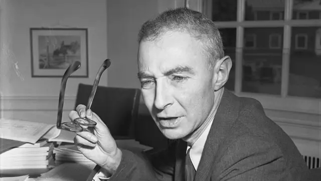 J. Robert Oppenheimer falando enquanto tira os óculoscomo funciona futebol virtual bet365frente a mesa com vários documentos