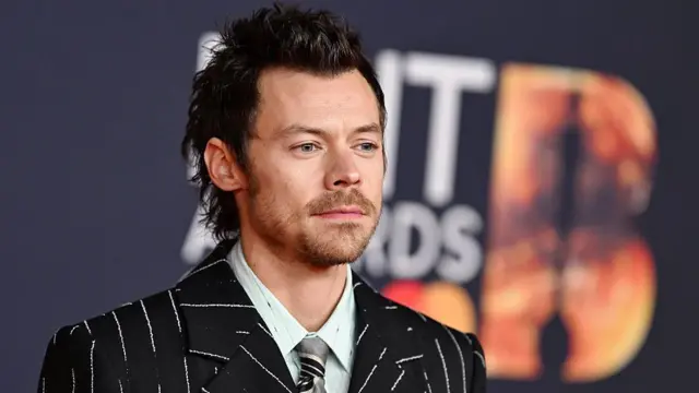 Harry Styles ma na sobie garnitur w prążki, pod którym widać jasnozieloną koszulę i srebrny krawat. Stoi na tle ścianki Brit Awards.