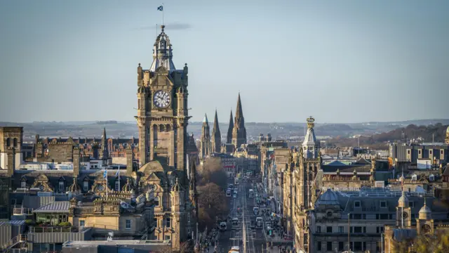 A imagem mostra uma vista panorâmica de Edimburgo, cidade marcada por edifícios históricos de pedra, com uma imponente torre de relógio dominando o horizonte. A avenida principal se estende em linha reta pelo centro da cena, cercada por construções de arquitetura vitoriana e neogótica, enquanto torres e pináculos se destacam ao fundo sob um céu claro.