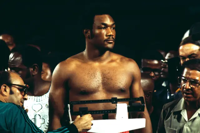 George Foreman is standing, shirtless, on a weighing scale during a weigh-in event. He is surrounded by several people, some partially visible, including a man operating the scale. The background is dark, with the crowd and event setting creating a focused atmosphere typical of a pre-fight weigh-in.