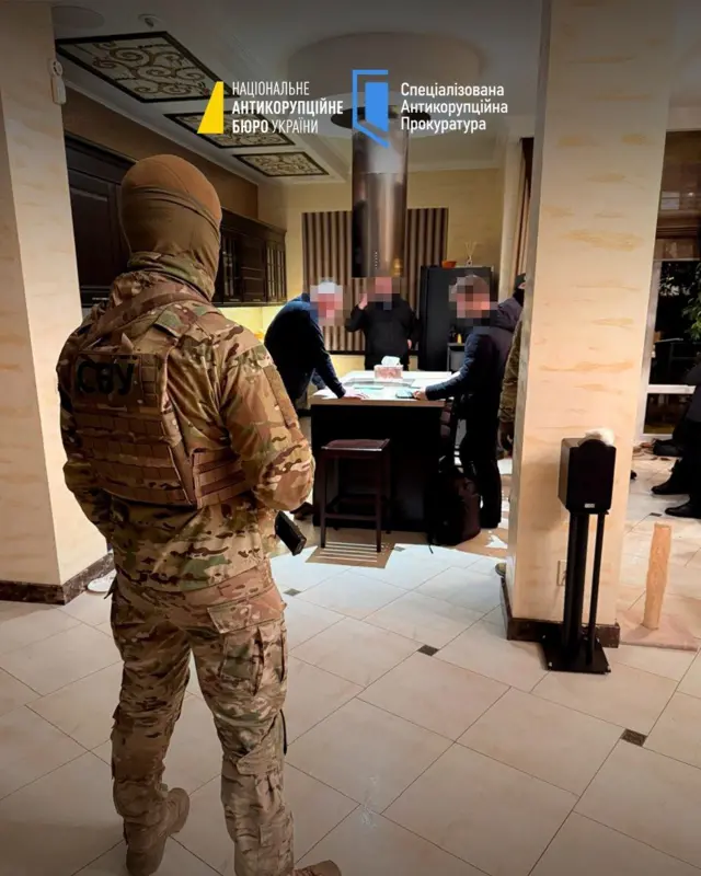 На передньому плані чоловік у формі в піксель, на задньому плані три чоловіка біля стола, їхні обличчя розмиті 