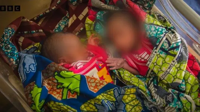 Awọn ibeji jojolo ti wọn fi asọ ankara alawọ eweko we, wọn tẹ wọn si ori ibusun, awọn ibeji naa wọ asọ alawọ pupa, ti a si fi nnkan bo oju wọn tori ọmọ kekere ni wọn.