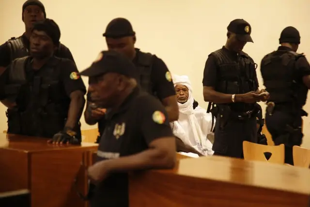 Habré, vestido con un largo boubou blanco, está sentado en una sala de un tribunal. Al frente de él, varios agentes de seguridad están parados