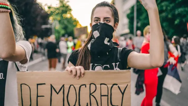Seorang perempuan muda pengunjuk rasa memegang poster bertuliskan "Demokrasi".