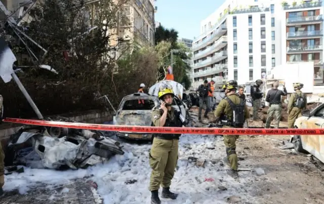 Tel Aviv mahallelerinden birini vuran füzenin yol açtığı hasar, yanmış araçlar ve haki üniformalı güvenlik görevlileri ve arka planda binalar