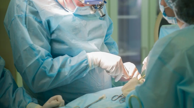 Médicos vestidos de azul, com máscaras e luvas brancas, realizando uma cirurgia