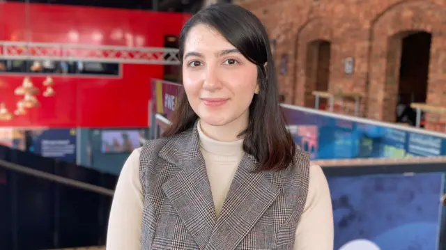 One young woman wit shoulder-length brown hair. She wear one smart double-breasted sleeveless jacket wit cream long-sleeve polo.