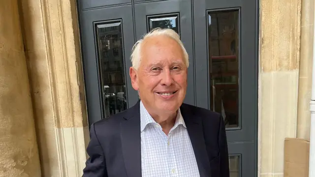 Sir Bob Neill está em frente a um edifício com grandes portas cinza-escuras e paredes de pedra bege. Ele está vestido com um paletó escuro sobre uma camisa xadrez azul-clara de gola aberta.