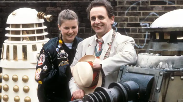 Sophie Aldred is wearing her black Ace jacket covered with badges, and is holding the arm of co-star Sylvester, who is in his iconic cream suit jacket. They are both smiling. Either side of them is an Imperial Dalek model and a Special Weapons Dalek.