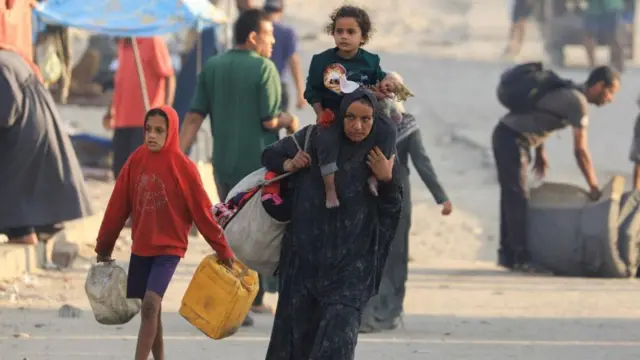 একটি মেয়েশিশু ও একটি বস্তা কাঁধে নিয়ে হেঁটে যাচ্ছেন এক নারী, তার পাশে একটি ছেলে শিশুর হাতে দুটি গ্যালন। পেছনে আরও কয়েকজনকে মালপত্র নিয়ে চলে যেতে দেখা যাচ্ছে