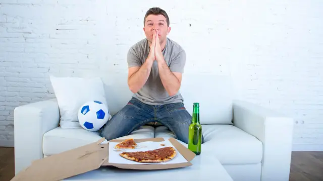 شاب يجلس على مقعد في وضعية مشاهدة مباراة على شاشة وأمامه قطعة بيتزا وزجاجة شراب وبجانبة كرة