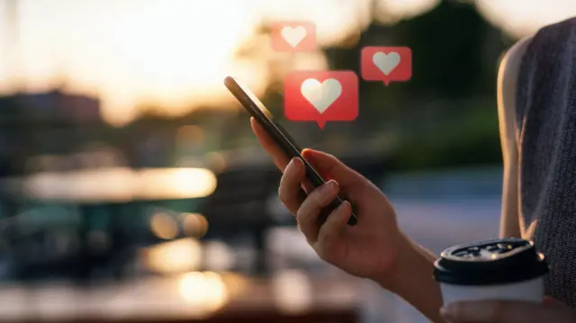 A photograph of a woman standing outside in soft evening light, background is not in focus. She is holding a phone in one hand and a paper coffee cup in the other. Love heart graphics are emerging from the phone. 