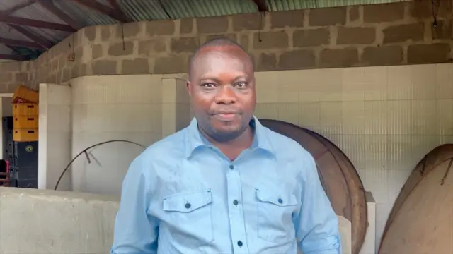 Umugabo wambaye ishati y'ubururu arimo kureba muri 'camera'. Inyuma ye hari urukuta n'igisenge cy'inzu. 