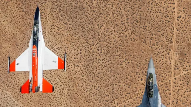 Two US Air Force F-16 combat jets fly side by side over the desert, one white and red, the other combat grey